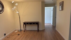 Um corredor com piso de madeira, um abajur, uma mesa de madeira escura com gavetas, um quadro na parede e uma porta branca no final.