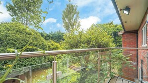 A balcony with a glass railing overlooking a garden with trees and a brick wall. - Willow Lodge 5,800 SQFT Quiet Luxury Living - Pass the Keys (Elstree)