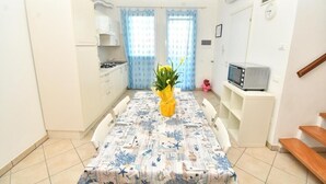 Una cucina compatta con mobili bianchi, un tavolo da pranzo con tovaglia fantasia, una piccola TV su un mobile e una pianta in vaso.