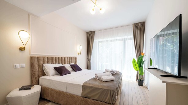 A modern hotel room with a large bed, a flat-screen TV, a bedside table, a potted plant, and a window with curtains. - Urban Amethyst Haven - 1BR Apartment with Balcony (București)