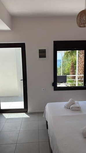 A bedroom with a bed, a window with a view of greenery and a building, and a door leading to an outdoor area.