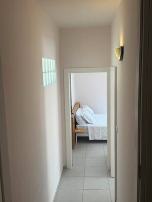 Un pasillo estrecho que conduce a un dormitorio con una cama, una mesita de noche y una lámpara fijada a la pared.