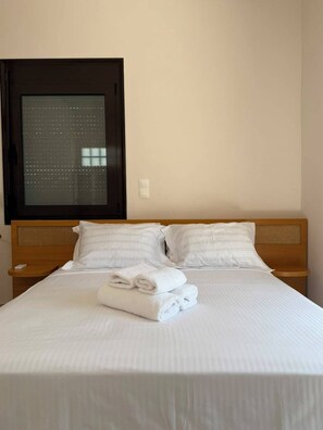 A neatly made bed with white linens and folded towels, a wooden headboard, and a window with blinds.