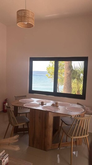 A dining area with a large window overlooking the sea, a wooden table, and chairs.