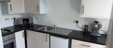 Una cocina con armarios blancos, encimera negra, horno empotrado, refrigerador, lavabo y cafetera.