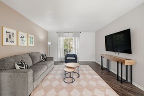 A modern living room with a grey sofa, a small round table, a TV mounted on the wall, and a window with curtains. - Winrock Apartments by Andes STR (Houston)