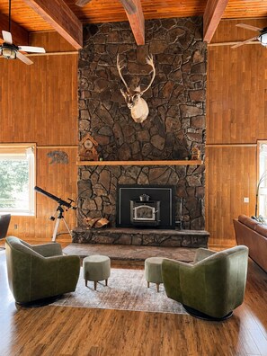 A room with a stone fireplace, wooden beams, and a mounted deer head. - Family-Friendly Cabin with Filtered Lake Views, Bunk Rooms, Game Space & Deck. (Cascade)
