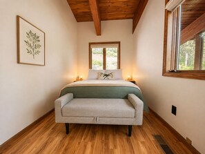 Une chambre à coucher avec un lit, des lampes de chevet, une fenêtre, une plante décorative dans un cadre et un plancher en bois.