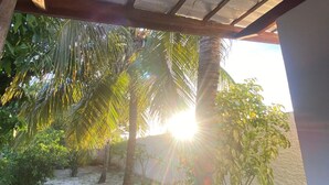 A covered outdoor area with a sandy ground, palm trees, and a view of the sun setting or rising. - House on the River (Barra do Jacuipe)