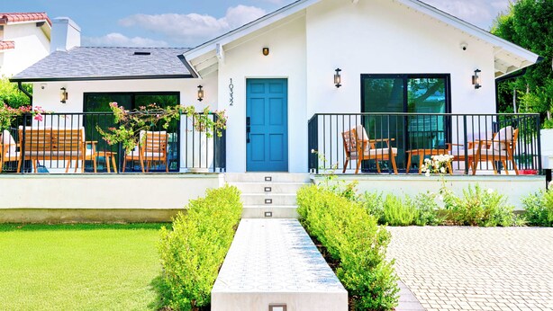 A modern two-story house with a blue door, a balcony with wooden furniture, and a well-maintained garden.