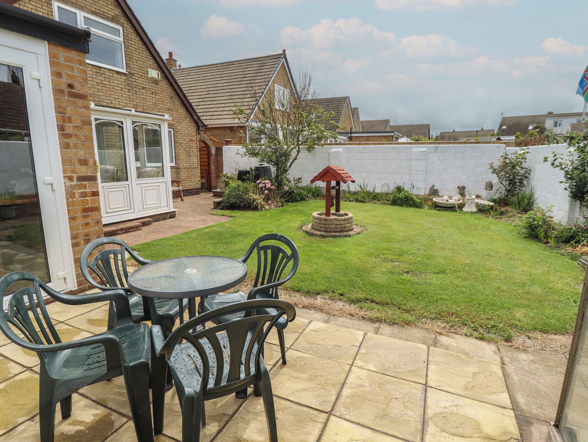 Um pátio com mesa e cadeiras, um pequeno jardim com um bebedouro para pássaros, e uma casa de tijolo com uma porta branca.