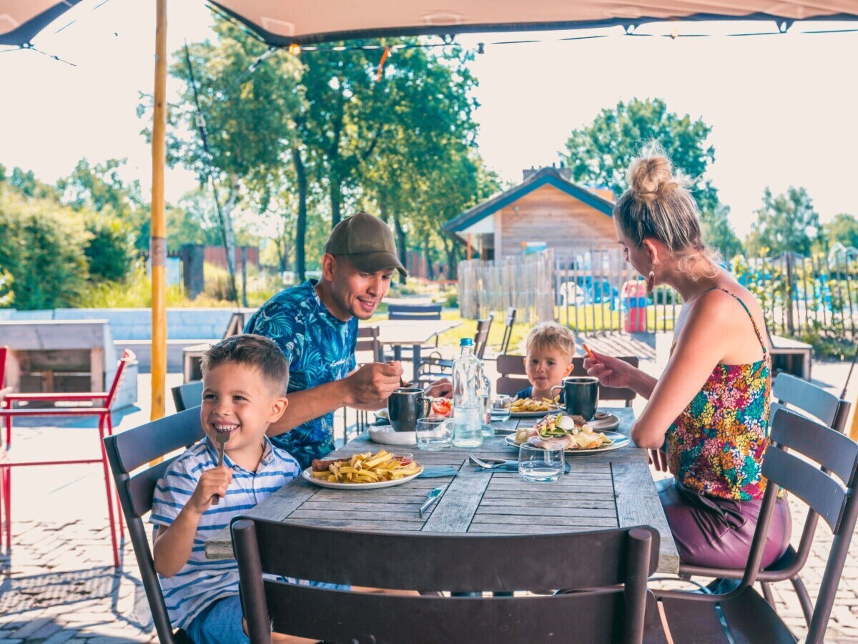 A family dining outdoors at a wooden table with chairs, a metal umbrella, and a house in the background.