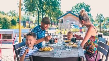 A family dining outdoors at a wooden table with chairs, a metal umbrella, and a house in the background.