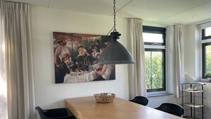 A dining area with a wooden table, black chairs, a hanging lamp, a painting on the wall, and a window with curtains. - Restyled Koningshoeve 4 People (Schoorl)