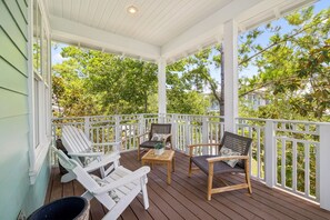Une véranda couverte avec des balustrades blanches, deux fauteuils Adirondack, une table en bois et un fauteuil en osier.