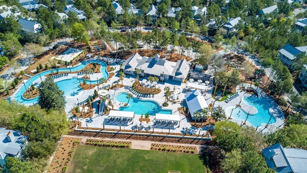 Eine Vogelperspektive zeigt einen Urlaubsort mit mehreren Schwimmbecken, einer weitläufigen Rasenfläche und angrenzenden Wohngebäuden.