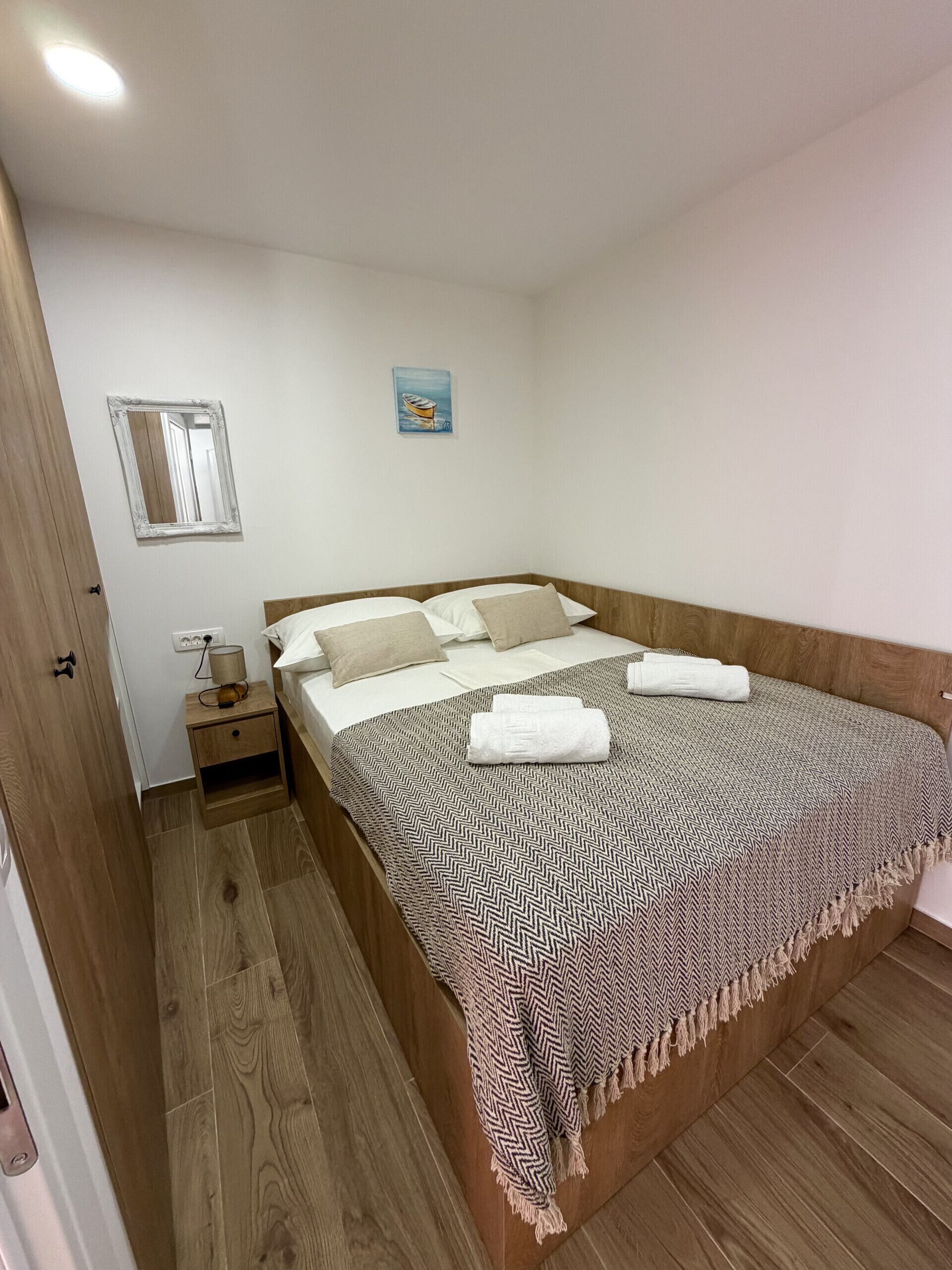 A bedroom with a wooden bed, a nightstand, a wall-mounted mirror, and a small painting.