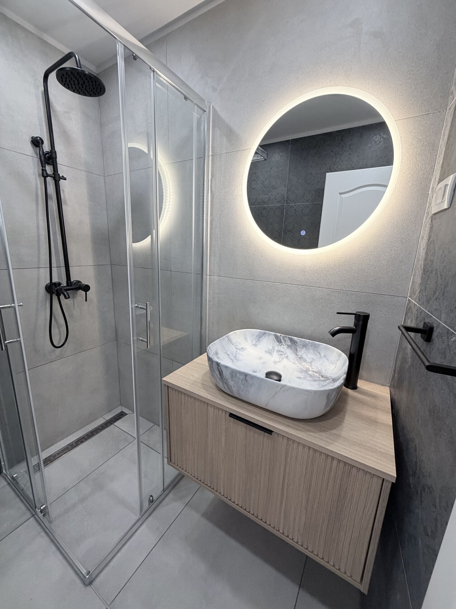 A modern bathroom with a glass-enclosed shower, a round mirror, and a marble sink.