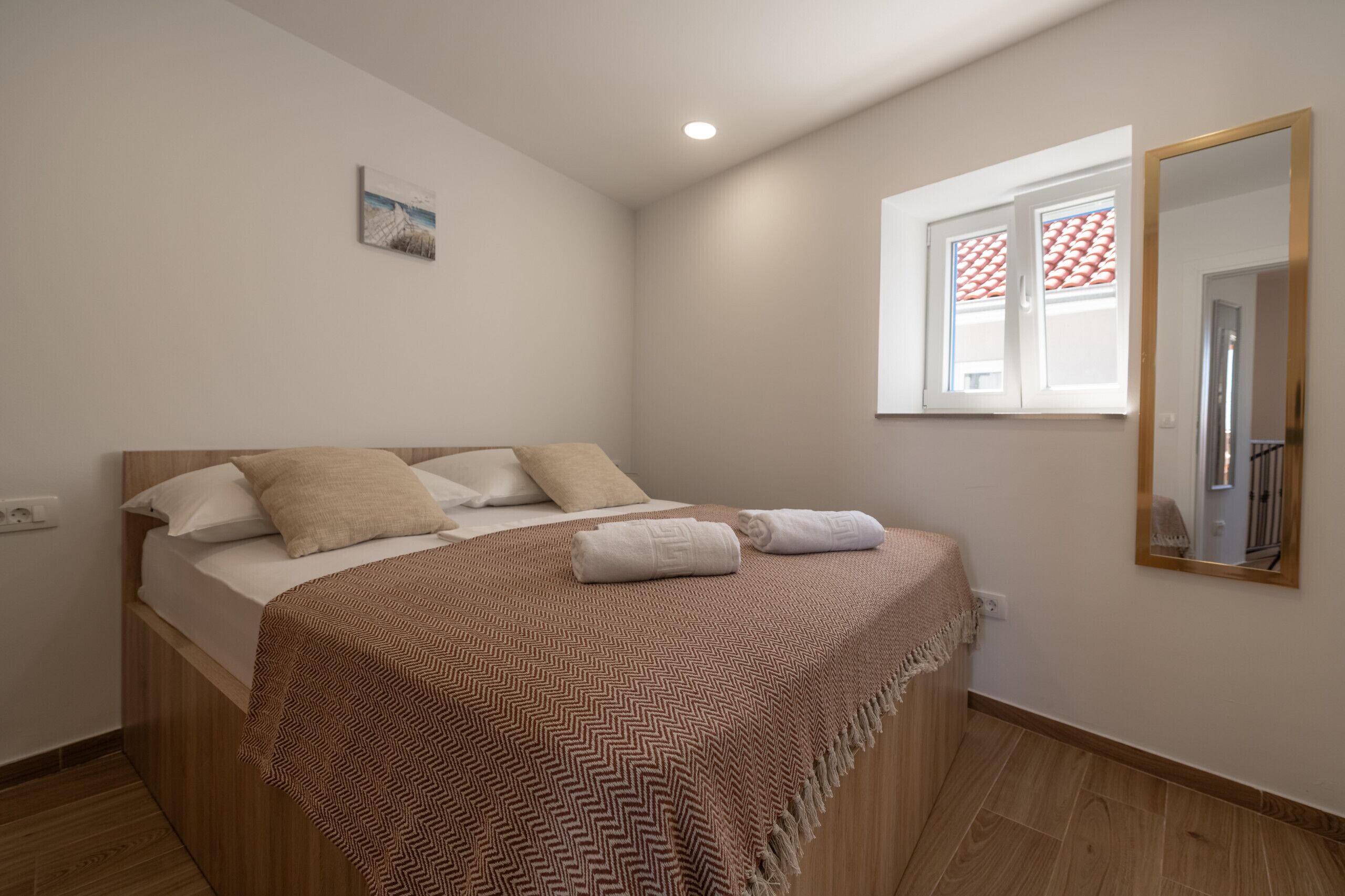 A bedroom with a bed, a window with blinds, a wall-mounted mirror, and a small picture.