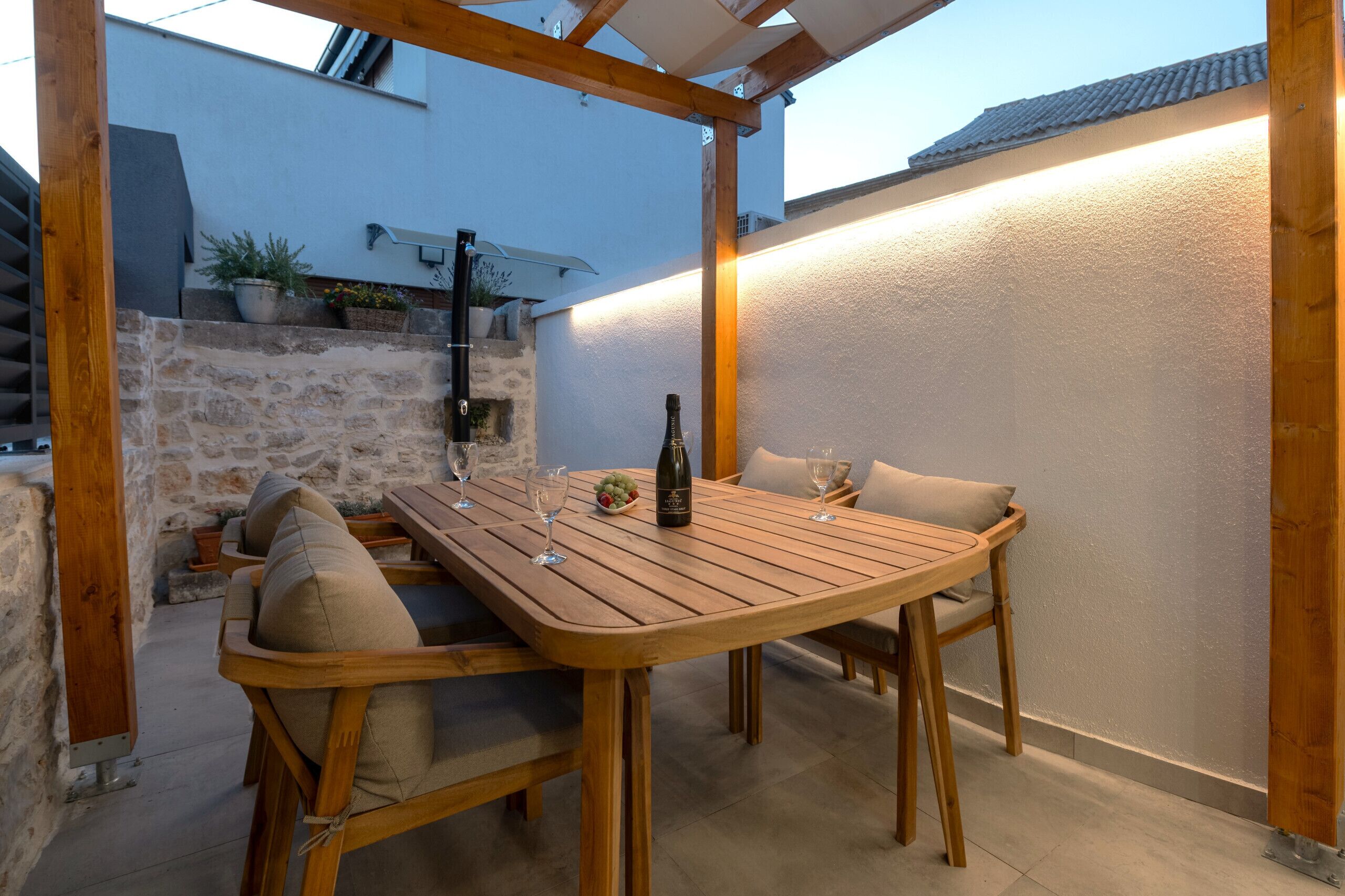 Een houten tafel met stoelen, een fles wijn en een schaal fruit op een terras met stenen muren en een pergola.