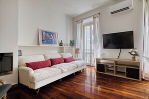 A living room with a white sofa, a flat-screen TV, wooden flooring, a window with curtains, and a wall-mounted air conditioner. - Garibaldi 44 - Milan Most Vibrant District (Milano)