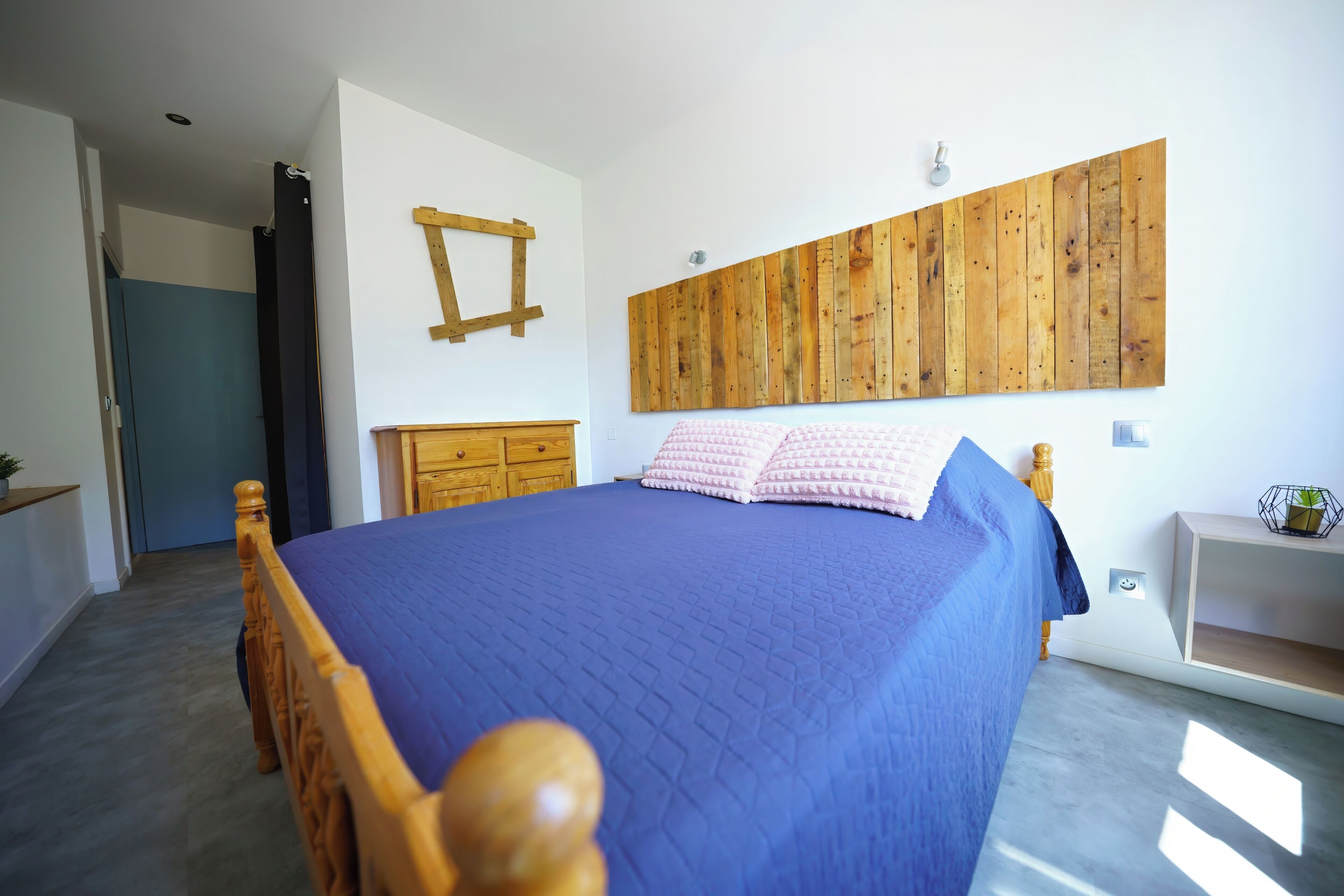 A bedroom with a wooden bed frame, a blue bedspread, a wooden headboard, a wooden wall panel, a wooden nightstand, and a small plant on the nightstand.