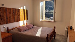 A bedroom with a wooden headboard, a bed with pink bedding, a window with a view of a hilly landscape, and a small potted plant on the nightstand.
