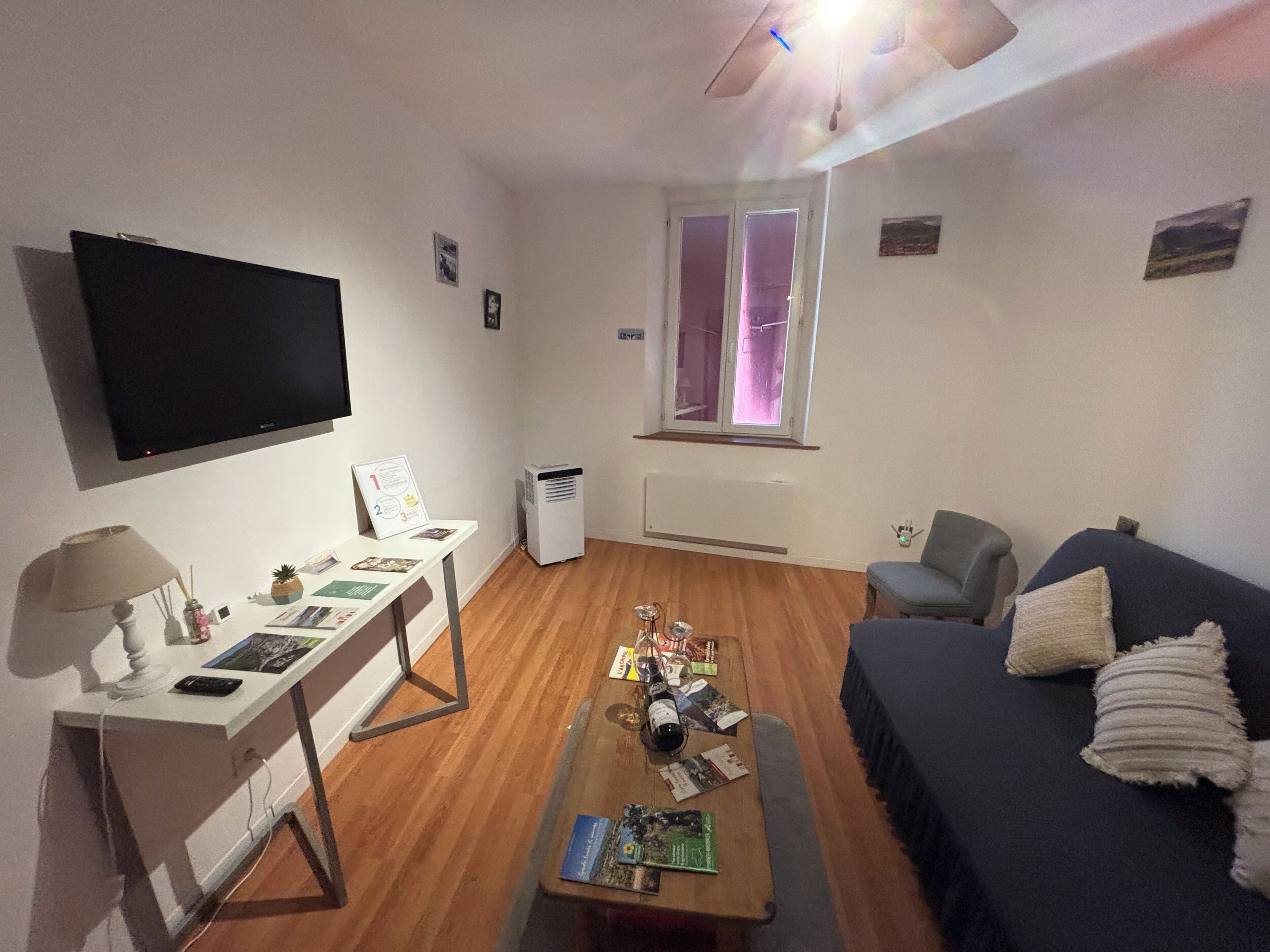 Un salon équipé d’un téléviseur à écran plat, d’un bureau avec une lampe, d’une table basse avec des magazines, d’un canapé et d’une fenêtre avec des stores.