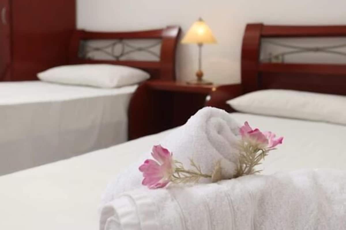 Habitación de hotel con dos camas, una mesita de noche con lámpara y una cama bien tendida con toalla y flores.