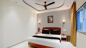 Una habitación de hotel con una cama, un ventilador de techo, una mesita de noche, una lámpara, una ventana con cortinas y un cuadro colgado en la pared.