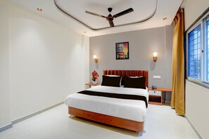 Une chambre d’hôtel avec un lit, un ventilateur de plafond, une table de chevet, une lampe, une fenêtre avec des rideaux et un tableau accroché au mur.