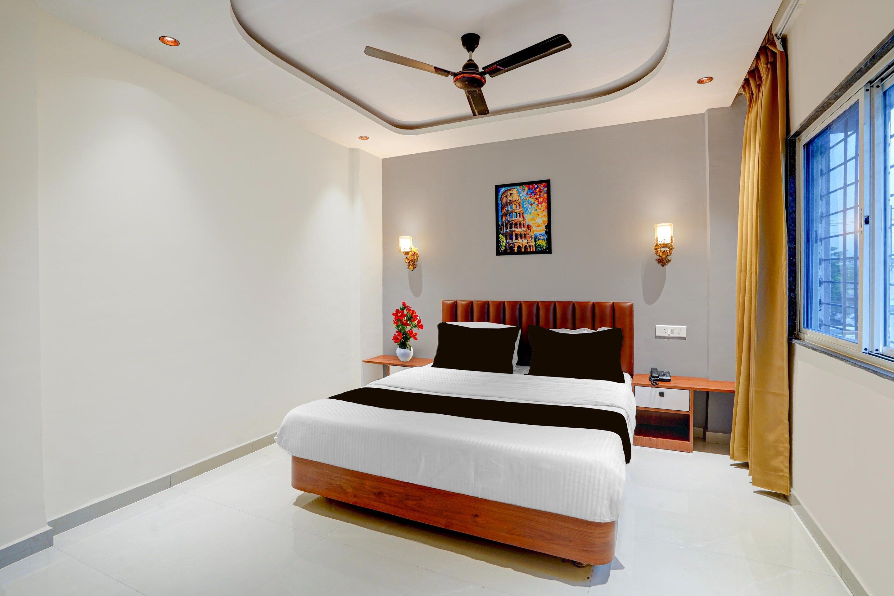 A hotel room with a bed, a ceiling fan, a nightstand, a vase with flowers, and a window with curtains.