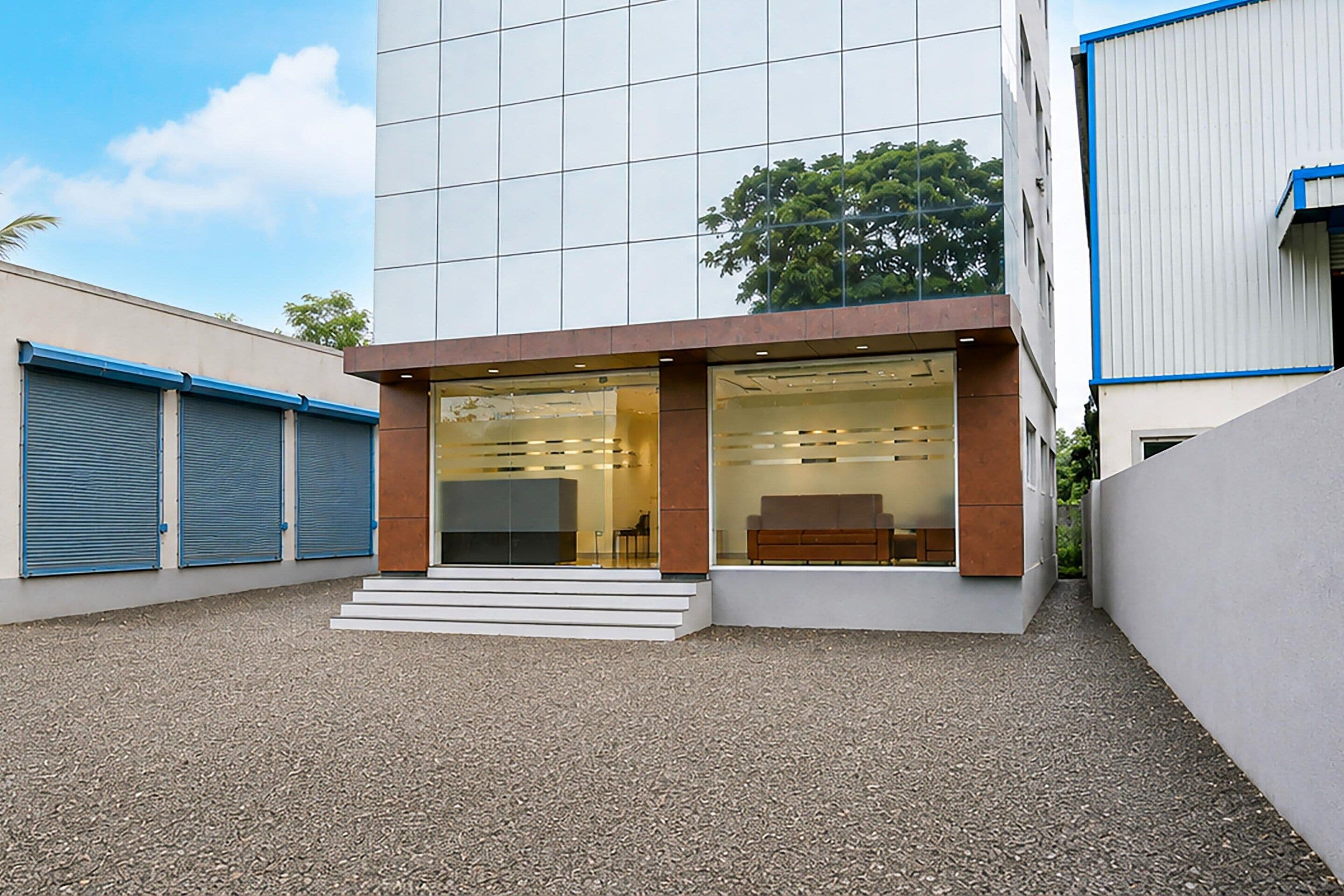 A modern building with a glass facade, a set of stairs leading to the entrance, and a parking area with gravel.