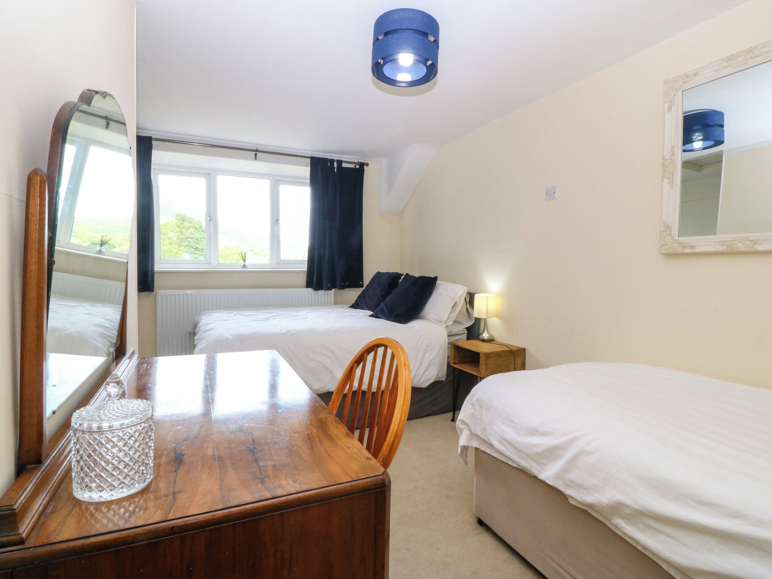 A hotel room with two beds, a wooden table with a glass container, a mirror, a lamp, and a window with curtains.