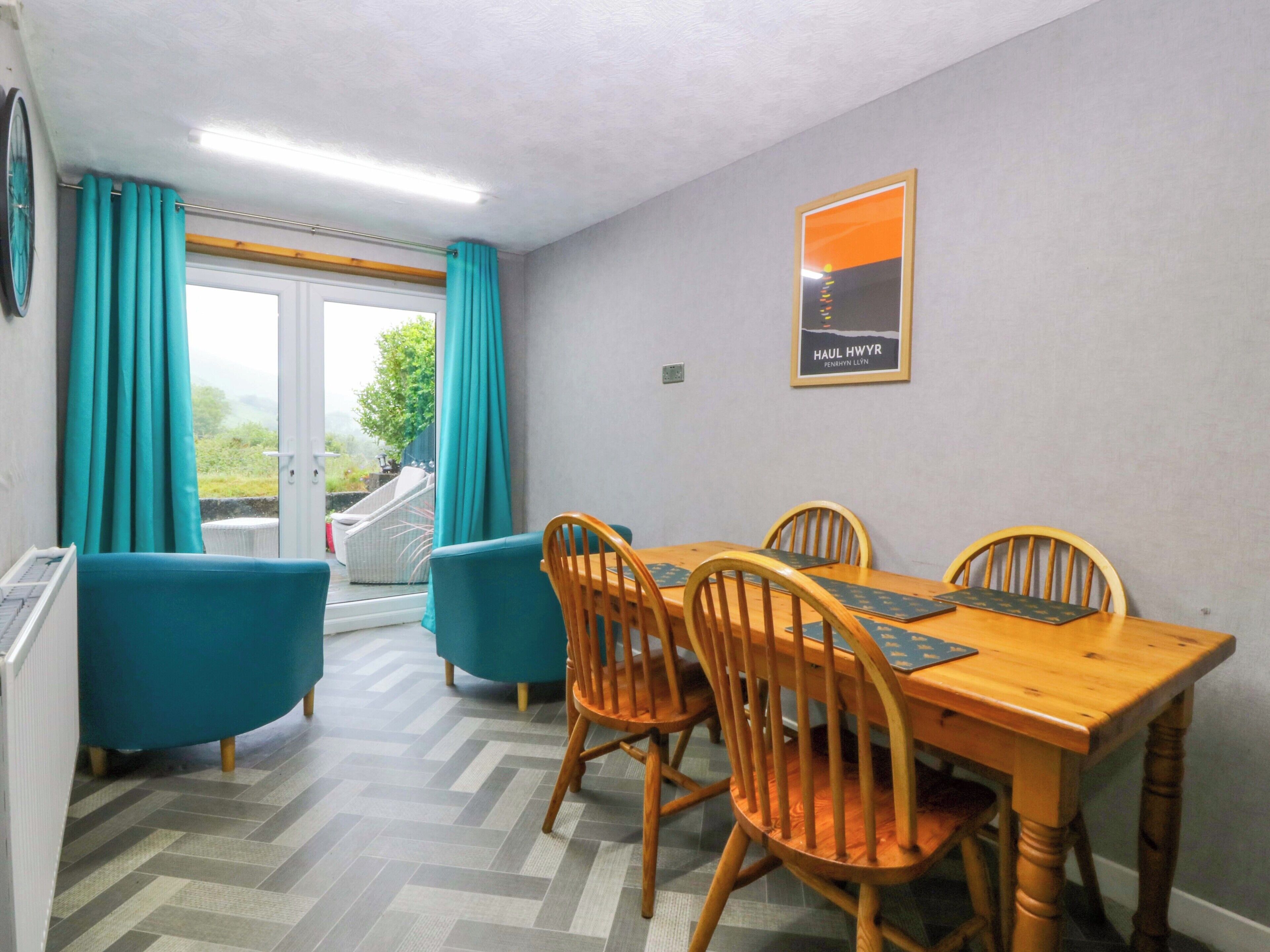 Uma sala de jantar com uma mesa e cadeiras de madeira, poltronas cor verde-água e vista através de uma porta de vidro.