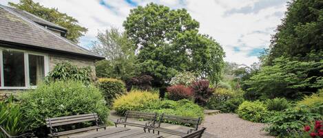 A garden patio with a table and benches, surrounded by various plants and trees.