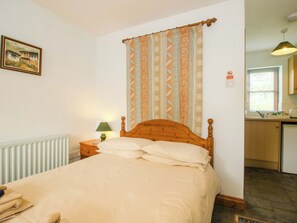 A bedroom with a wooden bed, a nightstand with a lamp, a radiator, a window with blinds, and a kitchen area visible through the door. - Apartment 2 (Lydbury North)