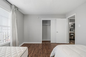 A bedroom with two beds, a closet, and a view of a cityscape through the window. - Wilcrest Apartments by Andes STR (Houston)