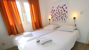 A hotel room with two beds, white linens, orange curtains, a floral painting, and wall-mounted lights. - Holiday Home in Breskens With Terrace (Breskens)