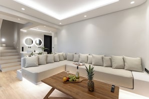 Ein modernes Wohnzimmer mit einer großen, L-förmigen Eckcouch, einem Couchtisch mit Früchten und einer Flasche sowie einer Treppe, die in eine obere Ebene führt.