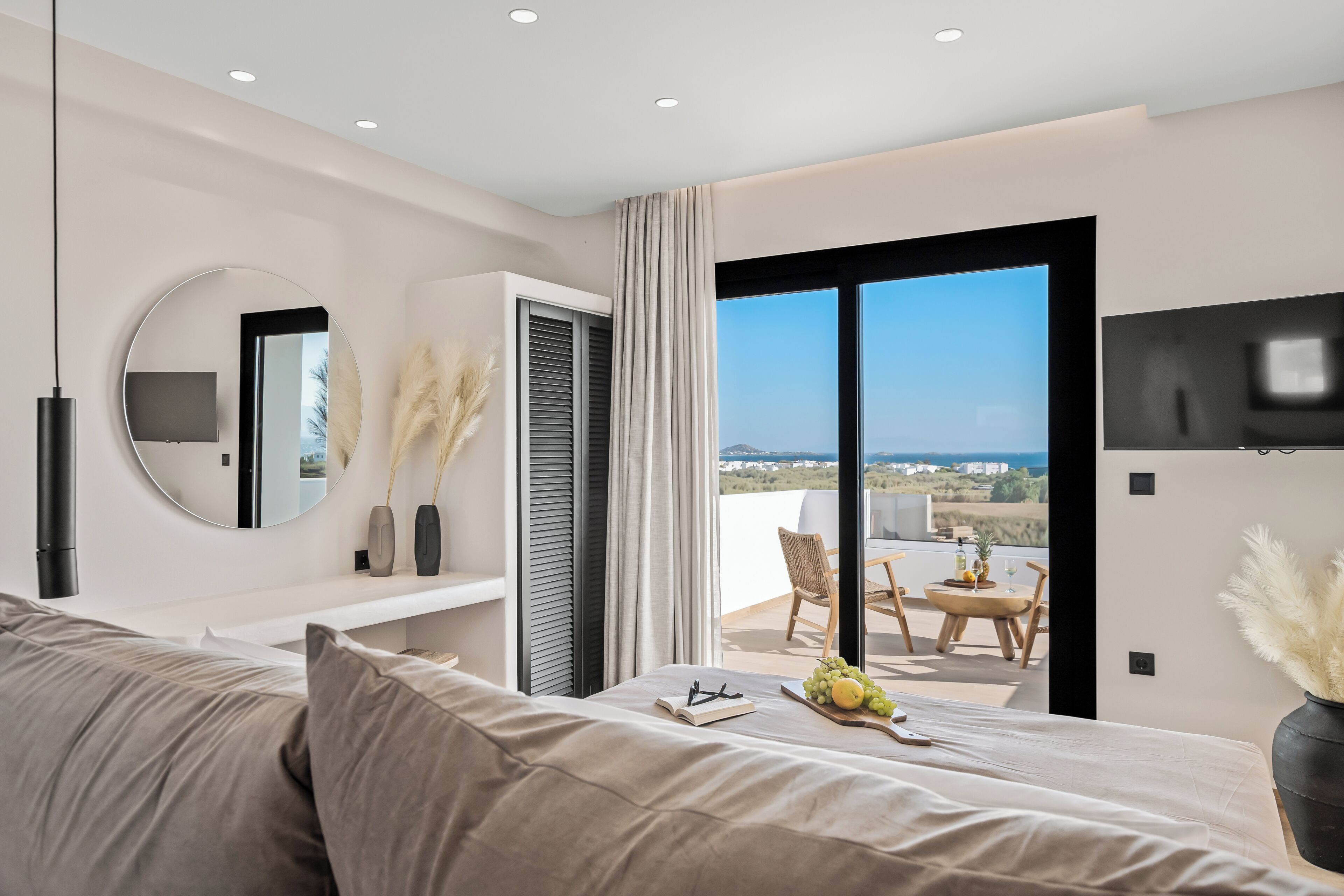 Quarto moderno com uma cama grande, um espelho redondo, uma TV e uma varanda com vista.