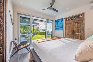 Ein Schlafzimmer mit einem großen Bett, einem Deckenventilator, Blick ins Freie und einer Holztür.