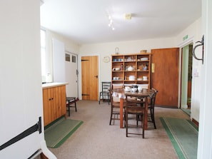 A kitchen with a dining table, chairs, a wooden cabinet, and a shelf with dishes. - Kwerky Cottage (Lydbury North)