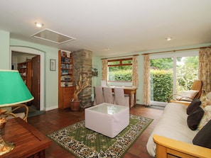 A room with a dining table, a sofa, a lamp, a bookshelf, and a view of greenery through the window. - Kwerky Cottage (Lydbury North)