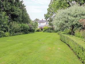 A well-maintained lawn with a hedge and trees, leading to a building with a white facade and a pitched roof. - Kwerky Cottage (Lydbury North)