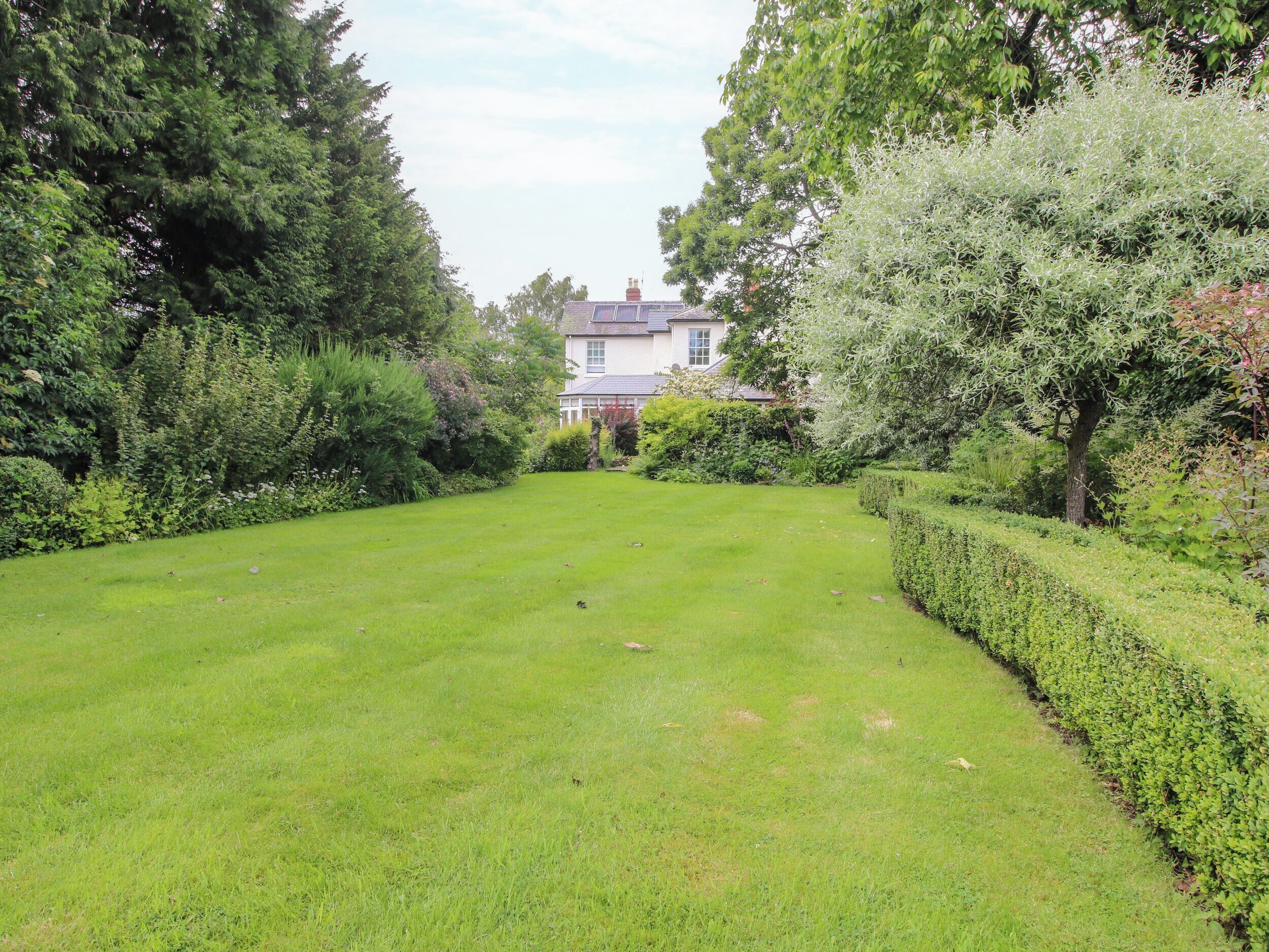 A well-maintained lawn with a hedge and trees, leading to a building with a white facade and a pitched roof.