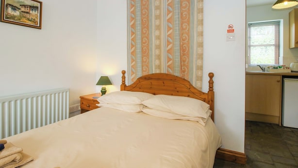 A bedroom with a wooden bed, a nightstand with a lamp, a radiator, a window with blinds, and a kitchen area visible through an open door. - Apartment 4 (Lydbury North)