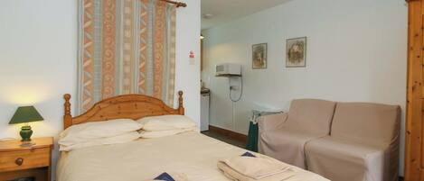 Un dormitorio con cama, mesita de noche con lámpara, una silla, escritorio con computadora y televisor montado en la pared.