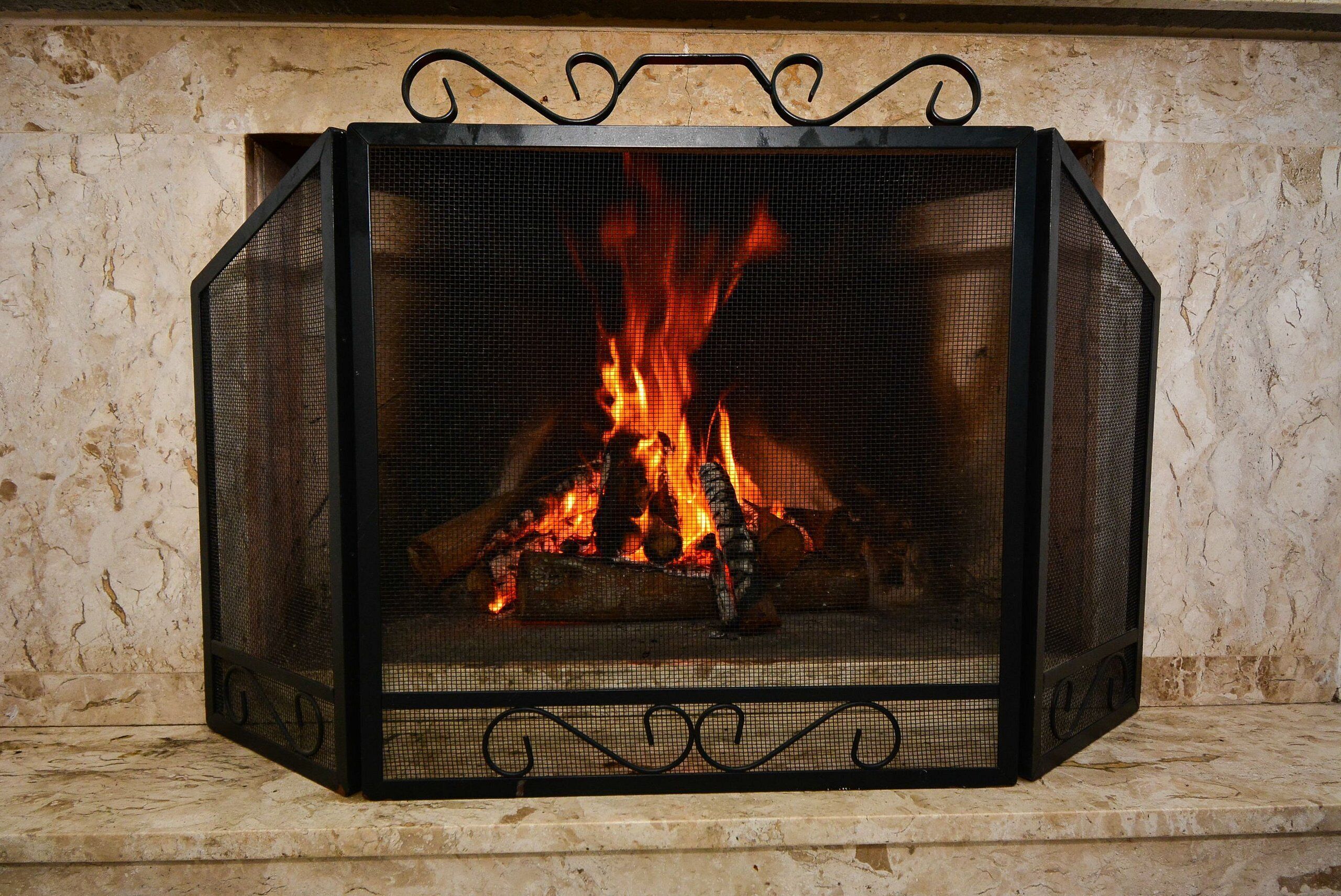 A fireplace with a metal screen, burning logs, and a marble wall.