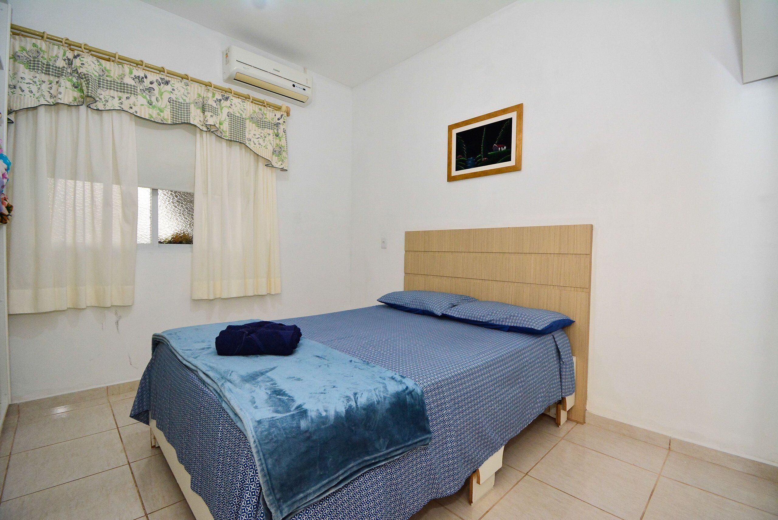 Quarto com cama, janela com cortinas, aparelho de ar condicionado e quadro na parede.